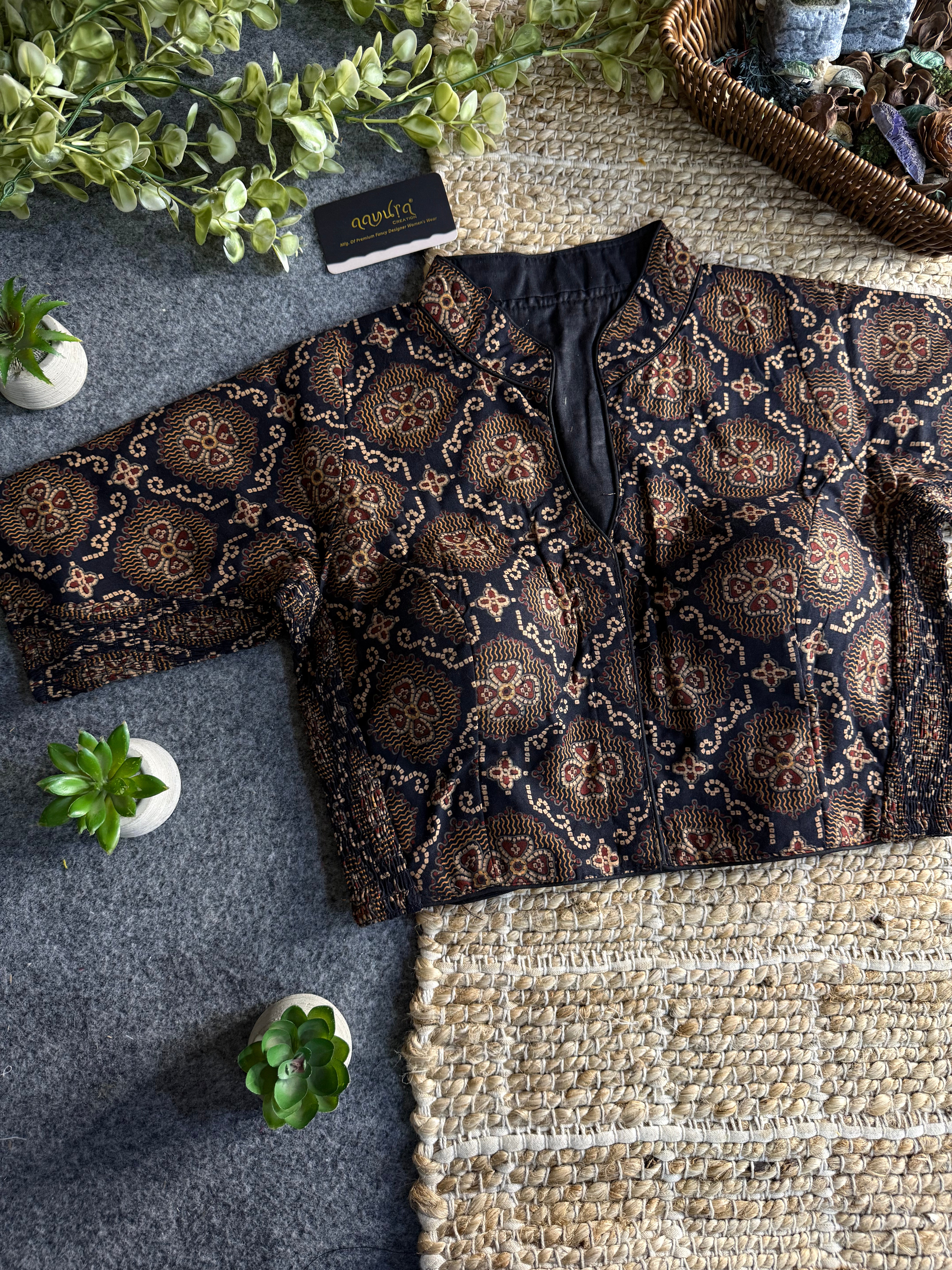 A cropped short-sleeve blouse with a black base and intricate brown and gold floral patterns, styled flat-lay on a gray textured surface next to a woven rug.
